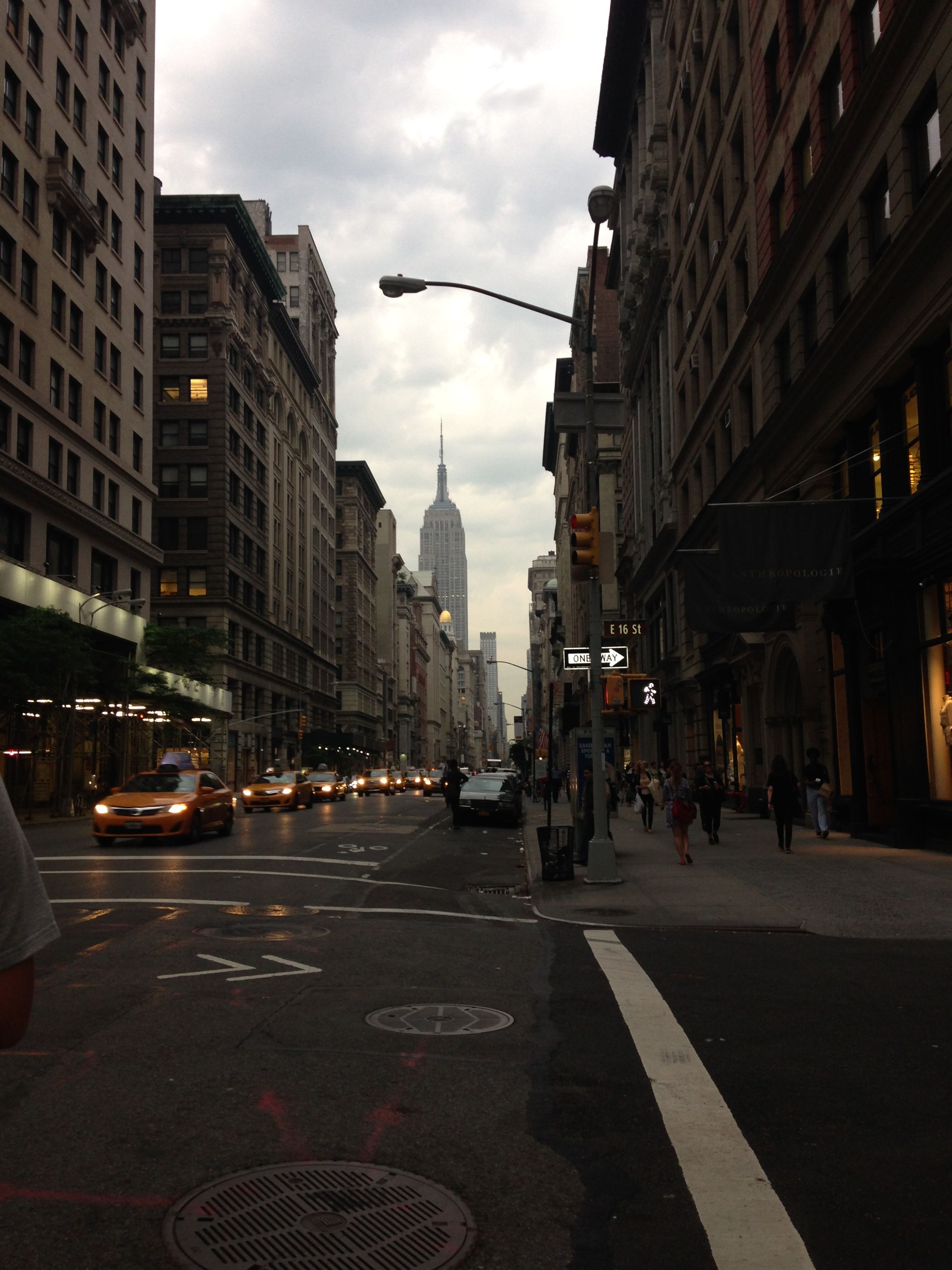 Photo taken on East 16th St in New York City with the Chrysler Building visible under a cloudy sky. A line of taxis is visible with their headlights on.