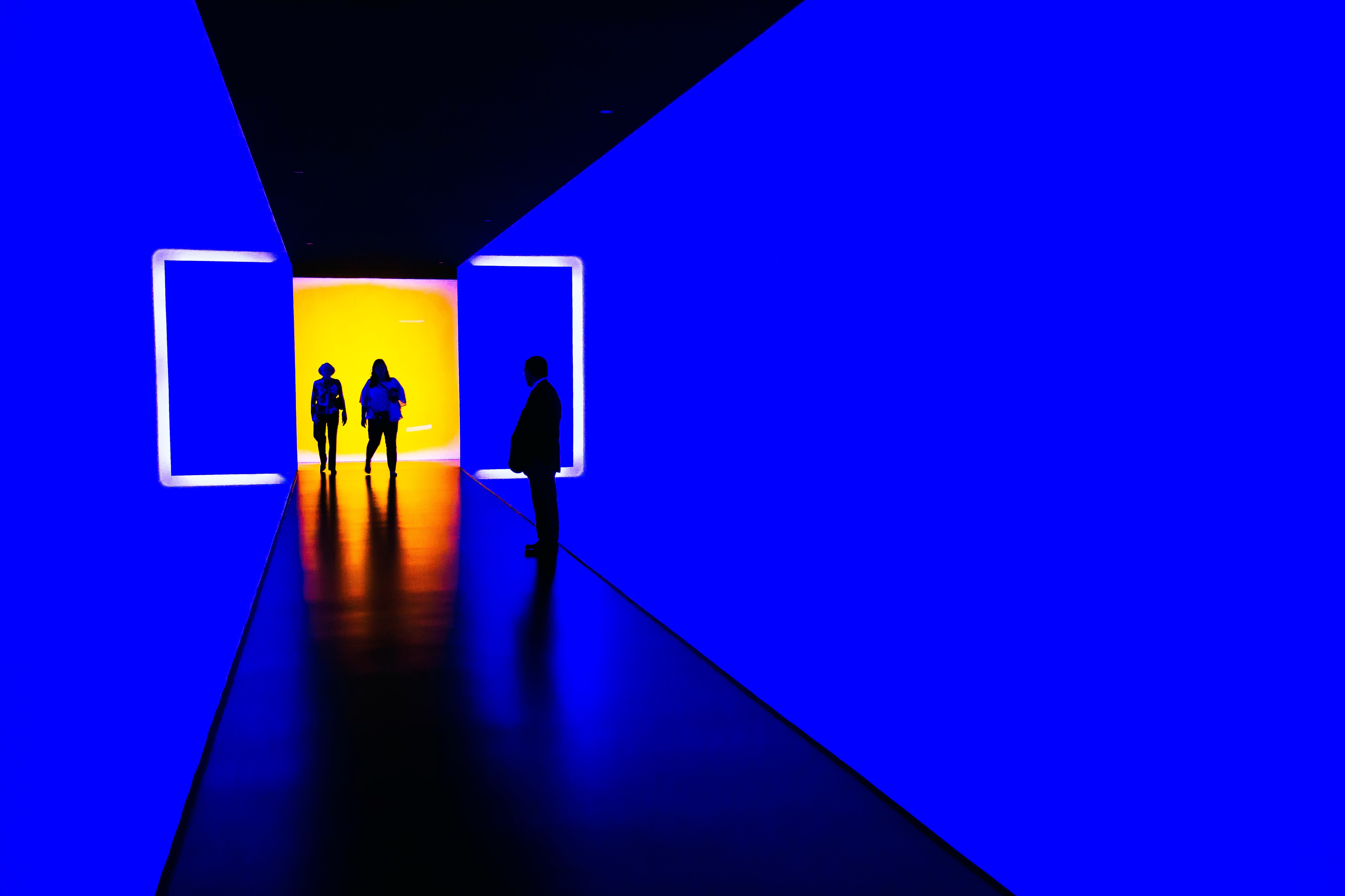 Three people standing in a hallway in a blue hallway in the Houston Museum of Fine Arts, Houston, TX. The room at the end of the hall appears yellow and frames two people walking in, while the third stands a few feet away turned toward them. The ceiling and floor appear black and the people appear framed by brackets of white light.